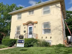 A two-story light yellow house undergoing exterior work, with plastic coverings taped over the windows and front door. The house has a small porch with a white railing and concrete steps. A "Malone" real estate sign is visible in the overgrown grass in the foreground.