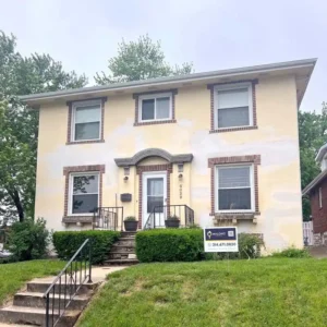 A light yellow, two-story house with a flat roof and brown brick trim around the windows and main entrance. The front of the house has a small porch with a white front door, a black railing, and concrete steps. A "Malone" real estate sign is visible in the lawn in front of the house.