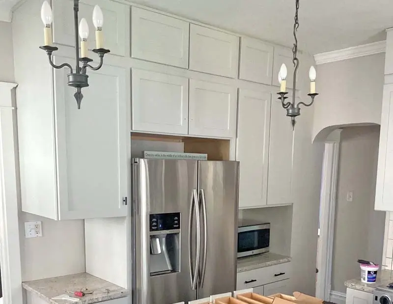 A stainless steel refrigerator and a microwave are surrounded by white cabinets in a kitchen. The room has light-colored countertops, and two chandeliers are hanging from the ceiling.