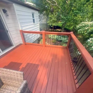 A view of a newly stained or painted reddish-brown wooden deck. The deck has a matching railing with black metal spindles and overlooks a small backyard with green plants and trees. A sliding glass door and the white siding of a house are visible on the left.
