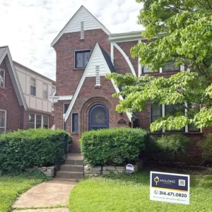 A brown brick, Tudor-style house with a large blue arched doorway and a second-story gable. The house has a stone retaining wall in front, with a neatly trimmed hedge and a lawn sign for "MALONE" with a phone number. The sky is overcast.