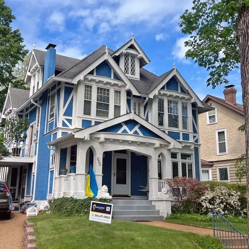 An outdoor shot of a two-story Victorian-style house with a blue exterior and white trim. The house has a complex roofline, a front porch with a white railing and columns, and multiple gables and dormers. Two white lion statues sit on either side of the front steps. A small blue and yellow flag is hanging on the porch railing. A "Malone" sign with a phone number is on the lawn in the foreground. The sky is partly cloudy and blue