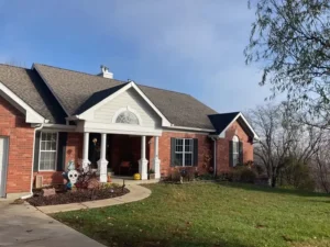 A ranch-style brick house with a brown roof and a covered front porch. The house has several windows with black shutters, and the front yard is decorated with Halloween-themed items, including a skeleton and a jack-o'-lantern. A concrete path curves from the driveway to the front door.
