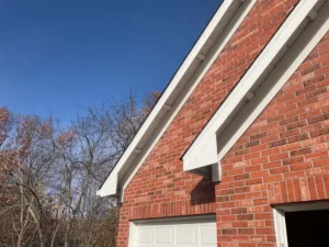 A low-angle close-up shot of the corner of a red brick house with a white garage door and white gutters. The clear blue sky is visible above the roofline. The bare branches of a tree can be seen on the left.