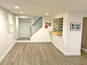 A newly finished basement area with light gray walls and wood-look flooring. The room features a bar area with a wooden countertop and wine bottle storage, and a staircase with a black metal railing leading to the upper floor. A white canvas and a framed photo are on the walls.