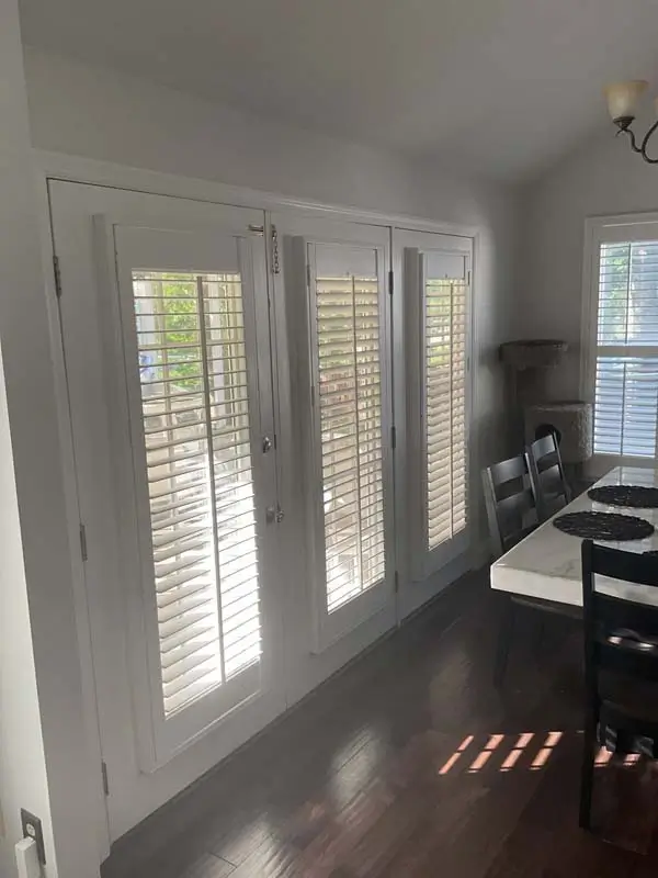 An indoor, horizontal shot of a dining area with dark wood floors. The image focuses on a set of white french doors with plantation shutters. Sunlight filters through the shutters and windows, creating striped patterns on the floor. To the right of the doors is a dining table with a white top and black chairs. A window with plantation shutters is also visible on the far right