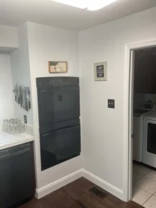 An indoor shot of a kitchen corner. The walls are a light color, and the floor is dark wood. A built-in black double oven is mounted in the corner. Above the oven, there are two small framed pictures. To the left, a counter with a white countertop holds several glass jars and a knife block is visible on the wall. A doorway on the right leads to a laundry room where a white washer and dryer can be seen