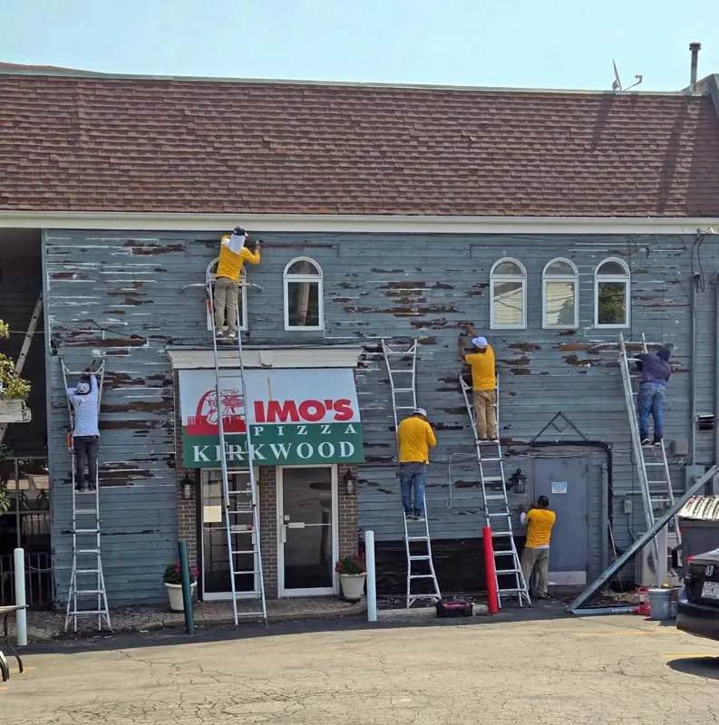A team of workers on ladders is preparing the exterior of a building for painting. The building has a sign for "IMO's PIZZA Kirkwood" and the siding is peeling. The workers are wearing yellow and white t-shirts. The sky is bright and clear.