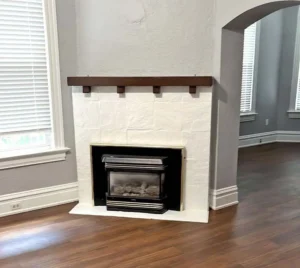 A modern fireplace with a white stone-like tile surround and a dark wood mantle. The fireplace insert is black and gold. The walls are a light gray, and the floor is a dark wood laminate. An arched doorway is visible to the right.