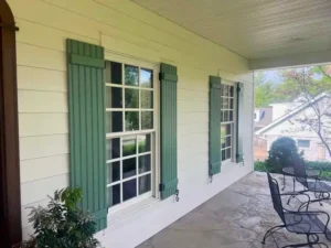 A close-up view of the front porch of a house with two windows, each flanked by decorative green shutters. The porch has a stone floor and a white ceiling with a single column visible on the right. A small table and two black metal chairs are on the porch.