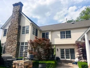 A side view of a large, light-colored house with a tall stone chimney on the left. The back of the house has numerous large windows and a set of sliding glass doors. An outdoor patio area is visible in the foreground with a grill, patio furniture, and a person kneeling on the ground near the house. A tree with reddish leaves is centered in front of the house.