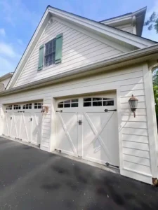 A low-angle view of a two-car garage with white doors that have a diagonal cross-brace design. The garage has light-colored siding, and there are two rustic-style wall lanterns flanking the doors. A single window with green shutters is visible on the upper level of the garage. The foreground shows a blacktop driveway.
