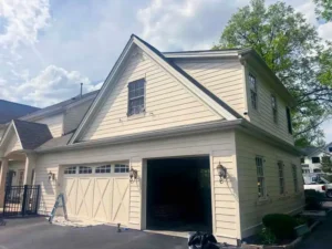A light-colored two-car garage with one white garage door and one open bay, revealing items inside. The garage has an attached second story with windows. A window with dark hardware is visible on the gable end of the garage roof.