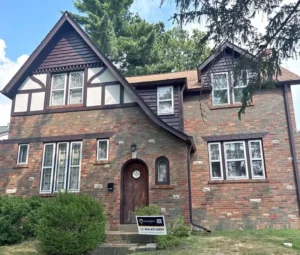 A Tudor-style house with a mix of red brick and brown-stained siding. The house has a wooden front door with an arched top, and a sign for "MALONE" with a phone number and QR code is in the front yard. The sky is partly cloudy, and trees are visible in the background.