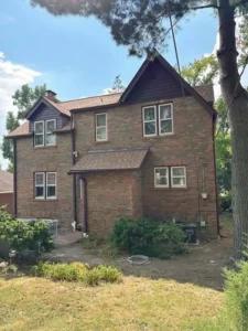 A large two-story brick house with a brown roof and matching siding on the upper dormers. The house has several white-trimmed windows and two air conditioning units on the left. A large tree trunk is visible on the far right, and the yard is a mixture of grass and dirt.