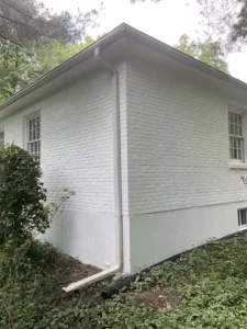 A white painted brick house with a concrete foundation. Two windows with white frames are visible on the side of the house. A white downspout runs along the corner, and the ground is covered in dark mulch and green plants, including ivy and a small bush. The roof is white and gray, and trees are visible in the background.