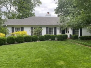 A ranch-style house with a white brick exterior and a gray shingled roof. The house has black shutters on the windows and a manicured lawn with a hedge and a few other plants. There is a white chimney on the roof, and the sky is overcast.