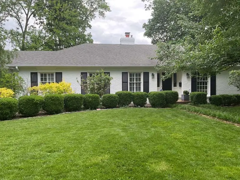 A ranch-style house with a white brick exterior and a gray shingled roof. The house has black shutters on the windows and a manicured lawn with a hedge and a few other plants. There is a white chimney on the roof, and the sky is overcast.