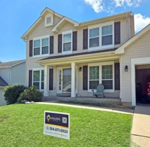An outdoor shot of a two-story suburban house with light tan siding. The house has a small front porch and a two-car garage on the right. A "Malone" sign with a phone number is prominently displayed on the front lawn. The windows have dark brown shutters. A light blue Adirondack chair is visible on the porch. The sky is blue with some white clouds