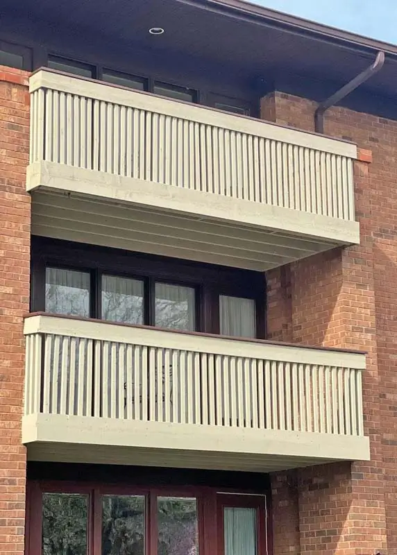 A two-tiered balcony with light-colored wooden railings on a red brick building. The windows and door below the balconies have dark brown frames.