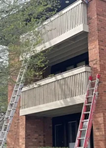 A side view of a red brick building with two light-colored wooden balconies. Two ladders are propped up against the building, one on the left and one on the right. The wooden railings on the lower balcony appear to be weathered and in need of repair.