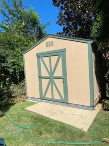 A tan-colored wooden shed with a green door that has a diagonal cross-brace design. The shed is on a concrete slab in a grassy yard, surrounded by green trees and bushes. A green water hose is lying on the grass in the foreground.