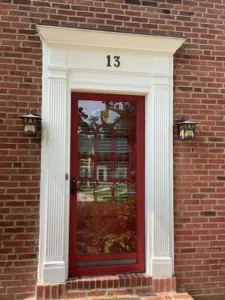 A close-up of a red storm door on a red brick house. The door is surrounded by a white frame with fluted columns and a pediment at the top, which has the number "13" on it. Two lantern-style light fixtures are mounted on the brick wall on either side of the door.