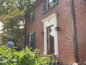 A red brick two-story house with white trim and dark green shutters on the windows. A white-trimmed front door with a red storm door is centered in the image. The number "13" is above the door. A purple hydrangea bush is in the foreground, and a large tree is on the left.