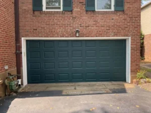 A dark green, paneled garage door with a white frame on a red brick house. A gas meter and a downspout are visible on the left, and a small flower bed is on the right. Two windows with closed green shutters are on the second floor above the garage door.