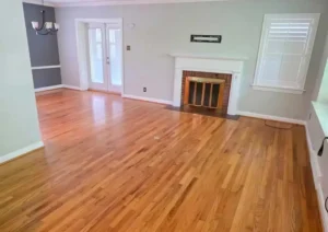 An empty living room with light hardwood floors. The walls are painted a light gray, with one accent wall painted a dark gray. A fireplace with a brick interior and a white mantle is centered on one wall. To the right is a window with white shutters, and to the left are a pair of french doors.