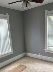 An empty room with light gray walls, white trim, and a beige carpet. A ceiling fan with a dark-brown blade and a four-light fixture hangs from the ceiling. A piece of wood is on the floor in the foreground, and there are two windows with white blinds on the left and right sides of the room.