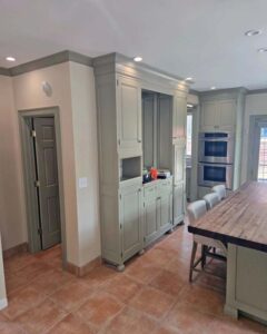 A large kitchen with sage green cabinets, a wooden island, and a tile floor. The cabinets extend to the ceiling and include built-in double ovens. A gray door is open on the left, and recessed lighting is visible in the ceiling.