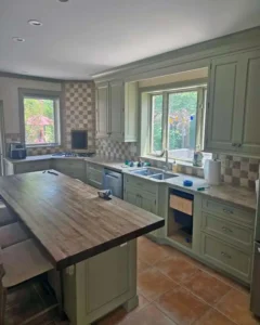 A kitchen with sage green cabinets, a double sink below a large window, and brown tiled floors. The countertops are a light speckled material, and the backsplash has a checkered pattern. A large wooden kitchen island is in the foreground, with tools and supplies visible on the counter.