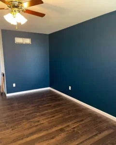 An empty room with dark wood-look flooring and navy blue walls. A gold and wood ceiling fan hangs from the ceiling. White baseboards line the walls, and a small, two-pane window is high up on the back wall.