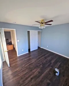 An empty bedroom with light blue walls and dark wood-look flooring. A gold and wood ceiling fan hangs from the ceiling. There is an open doorway on the left leading to a hallway, and a closet with white sliding doors in the back. A paint can is on the floor in the foreground, indicating the room has been recently painted.