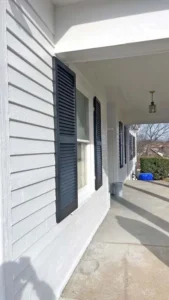 A view of a house's exterior siding, painted in a light gray color. There are multiple windows with louvered, dark gray shutters that are partially open. A concrete walkway and a covered porch are also visible. A blue recycling bin is in the background.