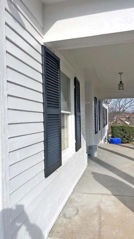 A view of a house's exterior siding, painted in a light gray color. There are multiple windows with louvered, dark gray shutters that are partially open. A concrete walkway and a covered porch are also visible. A blue recycling bin is in the background.