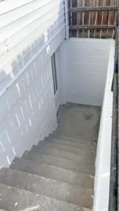 A view looking down a set of concrete stairs leading to a basement door. The walls on either side of the stairs are painted white, one made of brick and the other of cinder blocks. The basement door is partially visible at the bottom of the stairs.