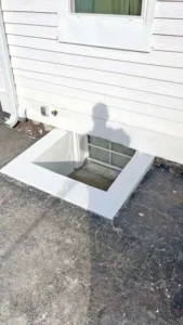 A white-sided house with a large square window well. The window well has a white frame and a glass-block window at the bottom. The ground around it is black and cracked. A shadow of a person is visible on the siding and the ground.