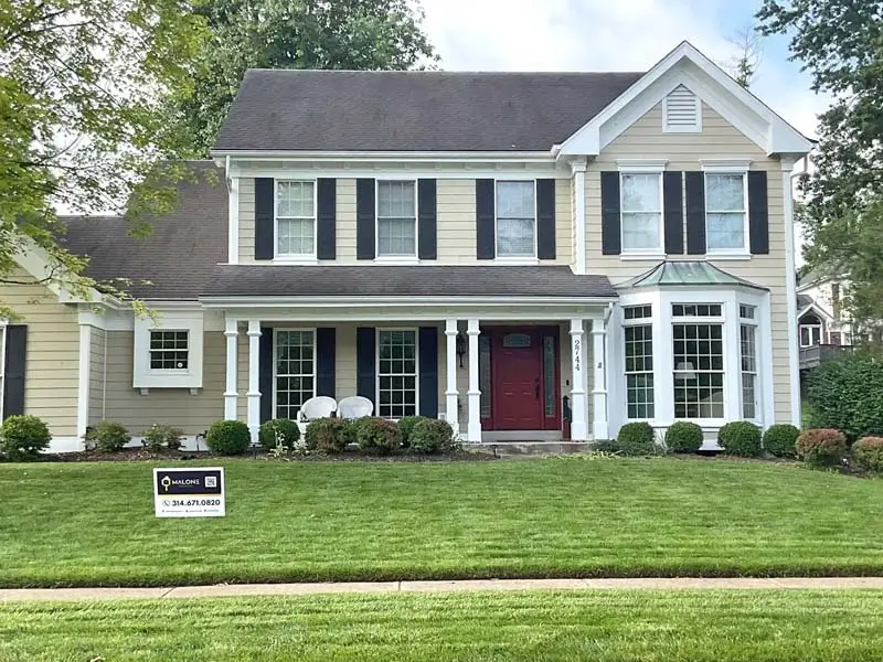 Front exterior of a two-story, light-yellow house with black shutters on the windows. A covered porch with white columns leads to a bright red front door, and a well-manicured lawn is in the foreground