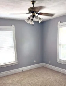 An empty room with light gray walls, white trim, and a beige carpet. A ceiling fan with a multi-light fixture hangs from the center of the ceiling. There are two windows with white blinds on the left and right sides of the room.