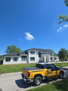 A low-angle, wide shot of a modern white house with a black roof and black-framed windows. A yellow and black Malone Painting company truck is parked in the driveway in front of the house. The sky is bright blue with a few white clouds.
