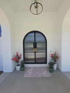 A view of the front entrance of a modern white brick house. The entrance features a large, arched black door with two gold handles and glass panels. A light-colored doormat is on the ground in front of the door. Potted plants with pink flowers are on either side of the door.