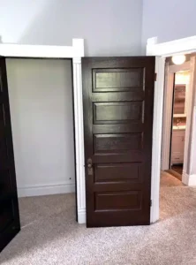 A brown five-panel wooden door standing open, revealing a closet. The door is propped against a white doorframe. A second doorway to a bathroom with a sink and mirror is visible to the right. The floor is covered with beige carpet.