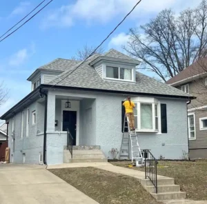 A light blue-gray stucco house with a gray shingle roof. A person in a yellow shirt is on a ladder next to a bay window, working on the side of the house. The front door is black and the windows have white trim. The yard is bare with brown grass, and a concrete driveway leads up to the house.
