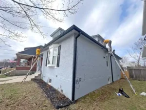 A two-story light gray stucco house with a dark gray roof. Two people wearing yellow shirts are on ladders, one on the front of the house and one on the side, appearing to be working on the gutters and roofline. The house has a bay window with black shutters, and the ground is covered in brown grass.