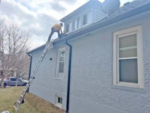 A two-story gray house with a stucco texture. A tall ladder is propped against the side of the house, and two people are on the roof near a dormer with multiple windows. A truck is parked on the street in the background.