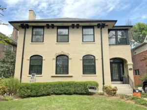 A two-story house with a light beige brick exterior and black window frames. The first floor has arched windows and an arched front door. A ladder is leaning against the house near the front door, and a small sign with the name "Malone" is on the lawn. A low hedge is in front of the house, and the sky is cloudy.