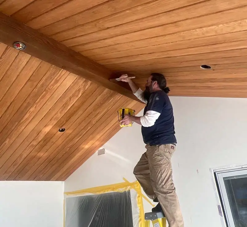 A bearded man on a ladder is painting a wooden, vaulted ceiling in a room. He is holding a paint bucket and a brush, and the surrounding walls are covered in yellow protective tape and plastic sheeting.