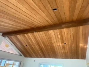 A low-angle view of a vaulted ceiling covered in light-colored wooden planks. A dark wooden beam runs across the center of the ceiling. There are several recessed light holes and a circular window at the peak of the roofline on the left.